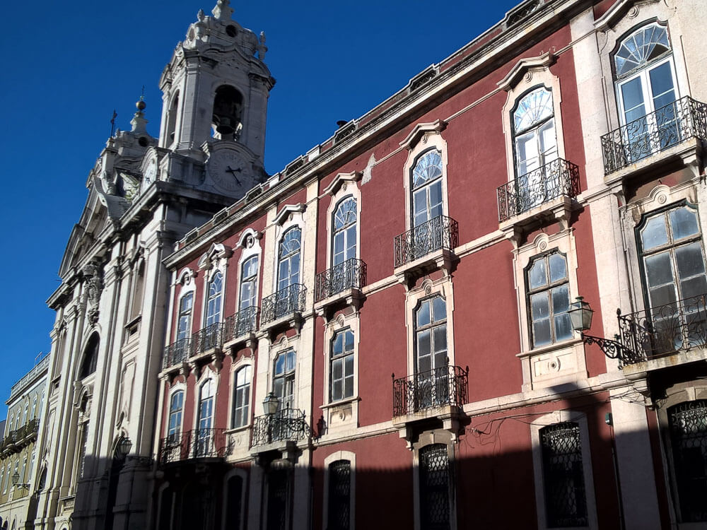 A view of the Igreja de São Francisco de Paula in Lisbon, with its red facade and ornate baroque architecture, including detailed balconies and a clock tower.