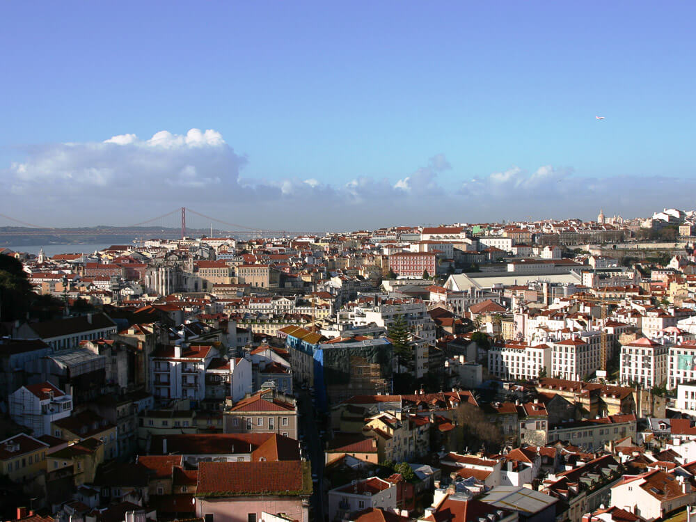 A wide view of Lisbon with red-roofed buildings and the 25 de Abril Bridge in the background.