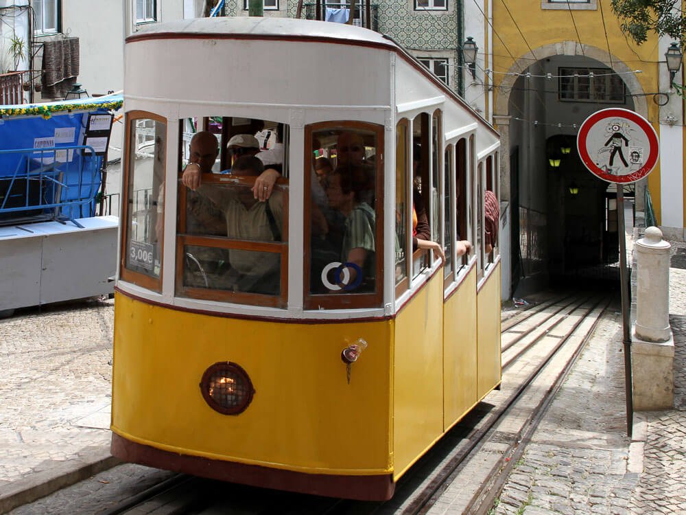 A yellow funicular with passengers inside, moving up a hill in Lisbon.