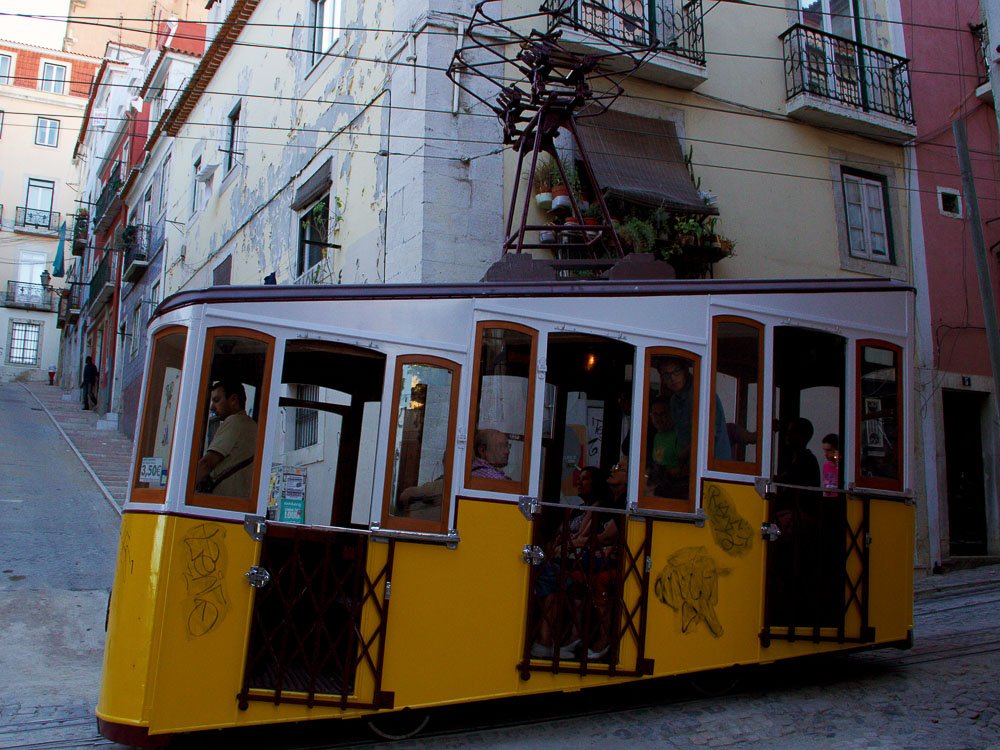 A yellow Bica funicular tram with passengers inside, traveling through a residential area in Lisbon.