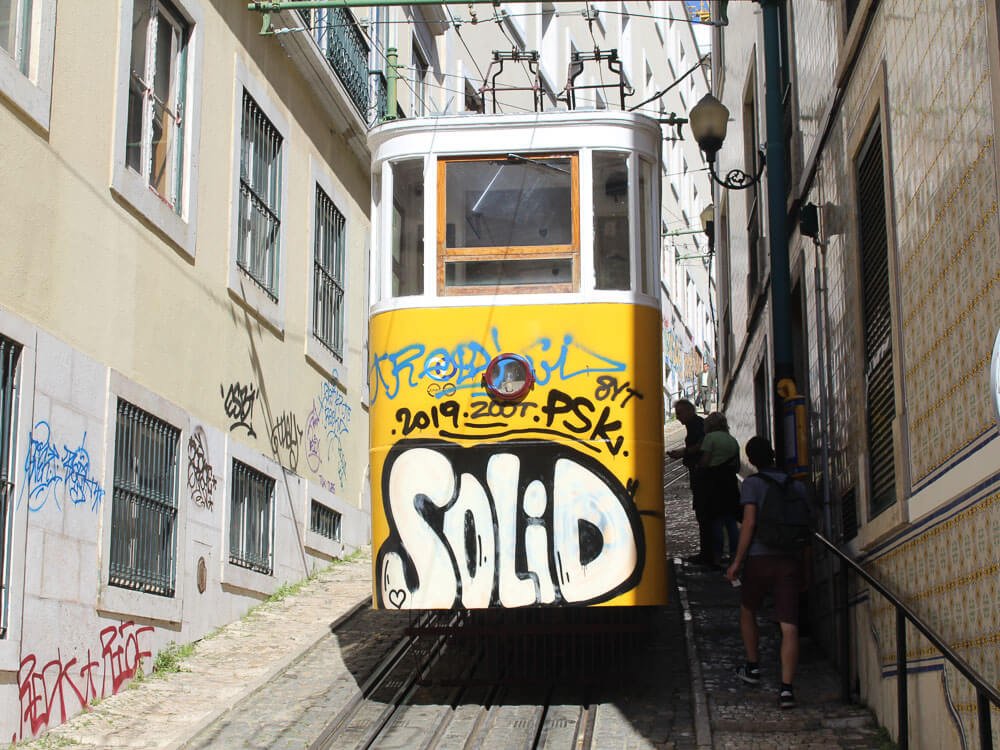 The back of a yellow funicular tram covered in graffiti, ascending a narrow, graffiti-lined street in Lisbon.