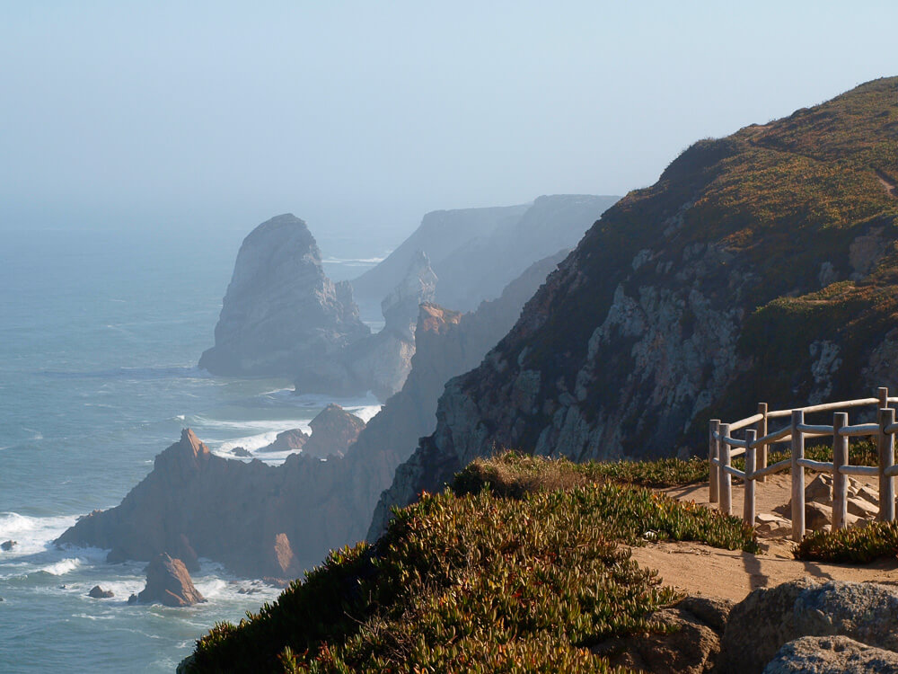 A stunning view of rocky cliffs in Cabo da Roca extending into the misty ocean, with a small viewing platform on the right.