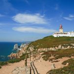 A wide-angle view of the cliffside path leading to the lighthouse in Cabo da Roca, with the ocean and distant cliffs in the background.