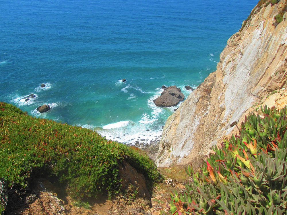 A steep cliff covered with greenery, leading down to the turquoise waters and rocky beach below.