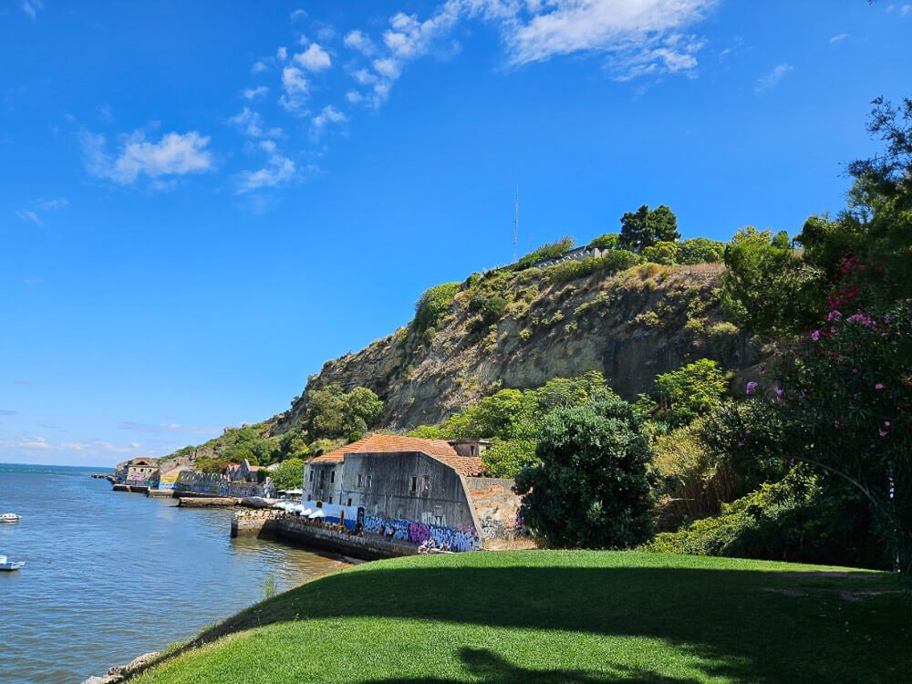 A serene coastal scene with green grassy areas, old buildings along the shore, and a blue sky.