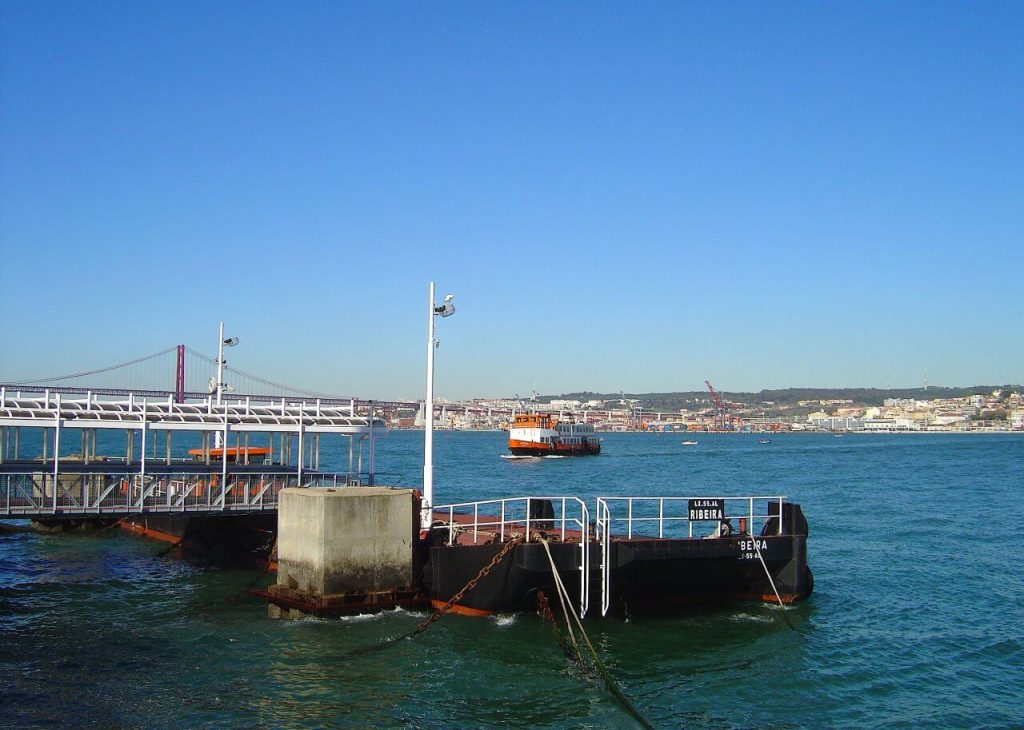  ferry dock with a boat approaching in the distance, set against the backdrop of the river with the 25 de Abril Bridge and the city visible on the horizon under a clear blue sky.