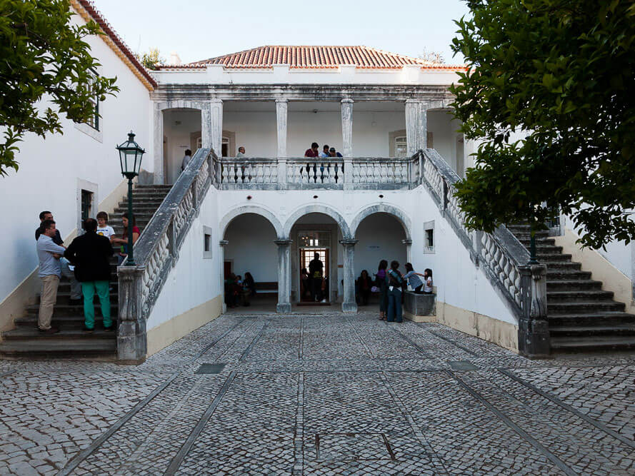 A courtyard of a historical building with an arched entrance, stairs on both sides, and people gathering in the area.