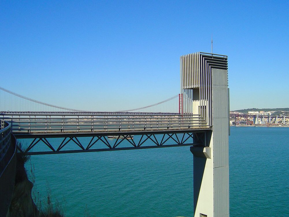 A section of a modern bridge extending over blue water, with the 25 de Abril Bridge visible in the background.