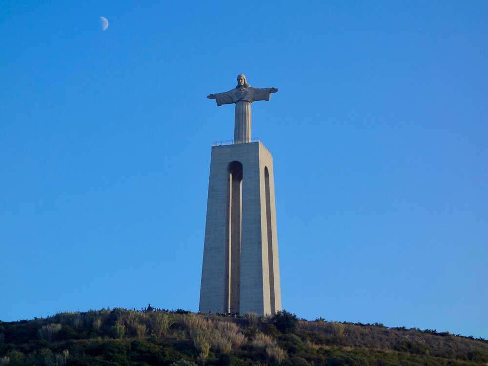 The towering Cristo Rei statue with outstretched arms, standing against a clear blue sky, with the moon visible in the background.