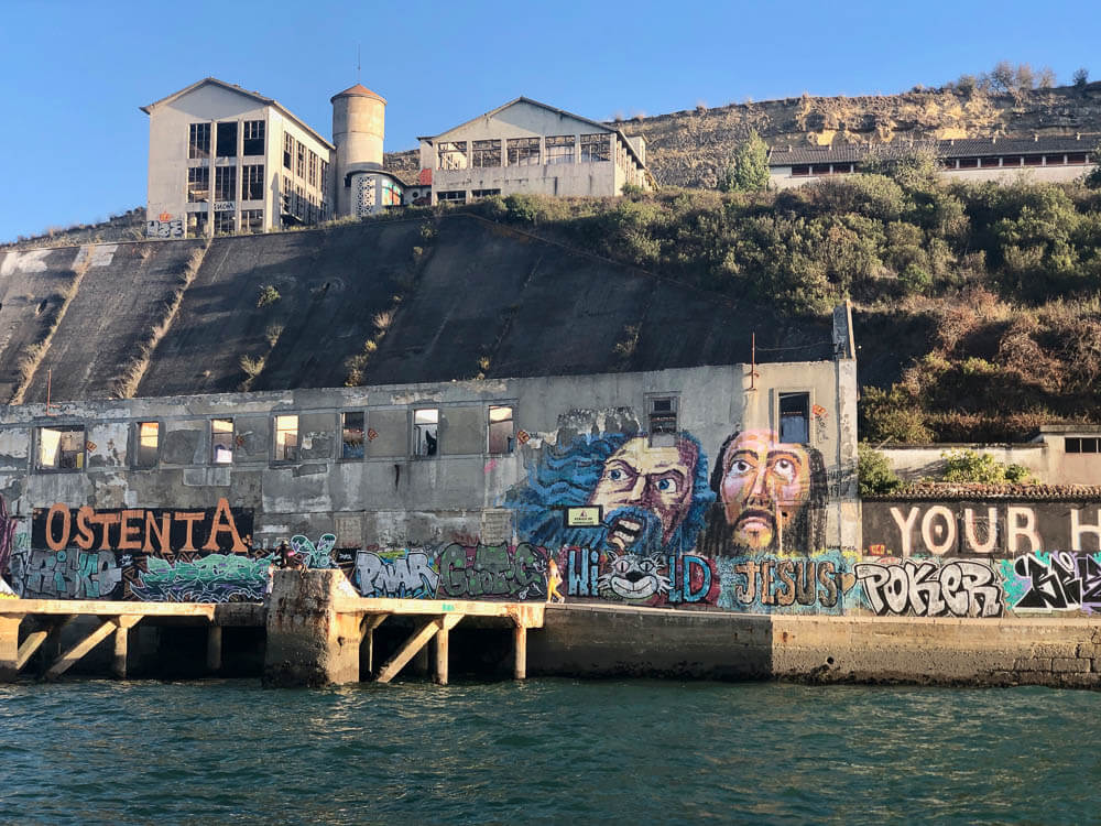 A derelict industrial building covered in colorful graffiti art, located on the waterfront.