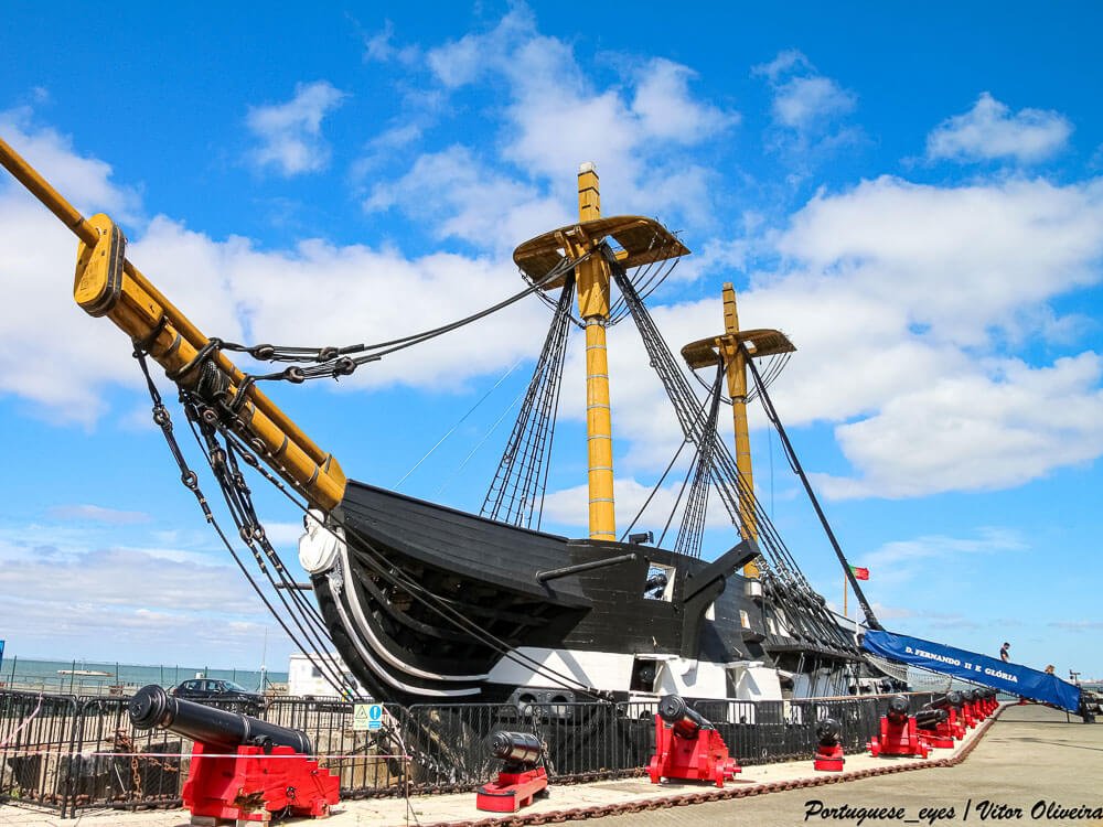 A close-up of a large, historical sailing ship with multiple masts, docked at a port under a blue sky.