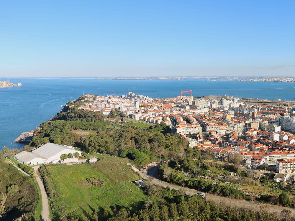 Aerial view of a coastal town with red-roofed buildings, greenery, and the ocean in the background.