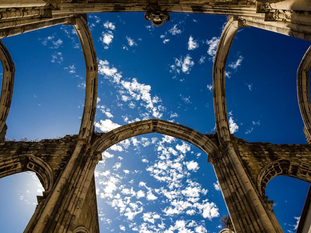 The arches of the Carmo Convent seen from below, with a backdrop of a bright blue sky and scattered clouds.