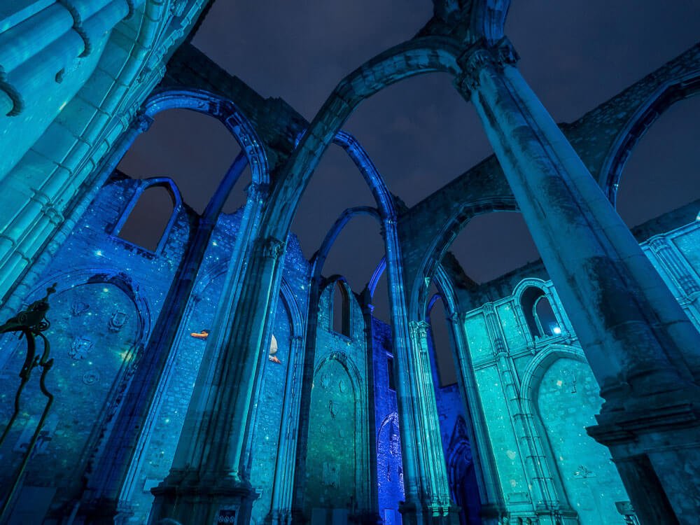 The Carmo Convent illuminated at night with blue lighting, highlighting the arches and walls.
