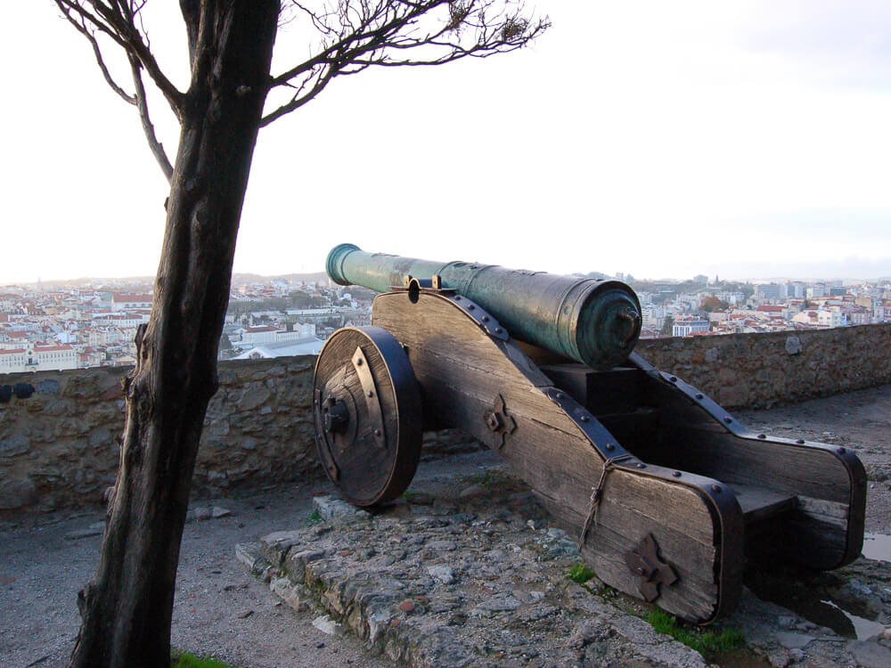 A cannon perched on the walls of Castelo de São Jorge, overlooking the city of Lisbon below, with a tree partially framing the scene.