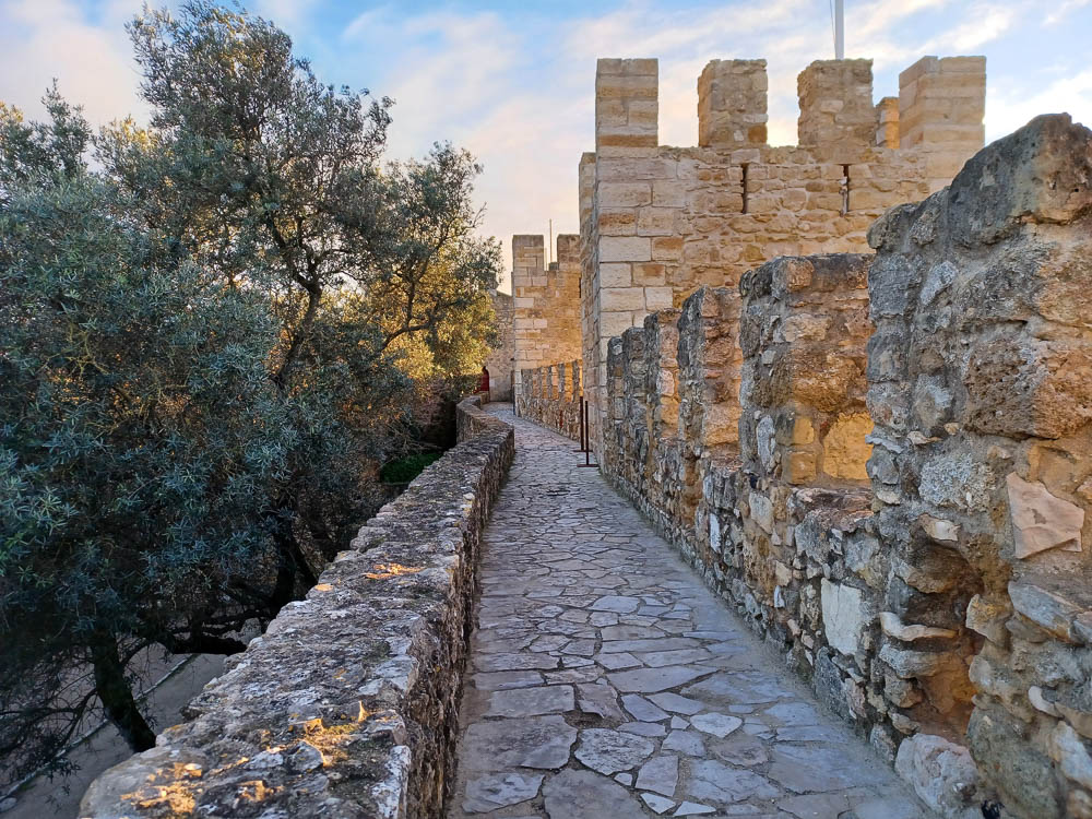 A narrow stone walkway along the fortified walls of Castelo de São Jorge, surrounded by trees and bathed in the warm light of sunset.