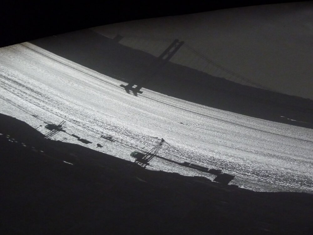 A black-and-white image of the Tagus River with a view of the 25th of April Bridge in the distance, taken from a high angle.