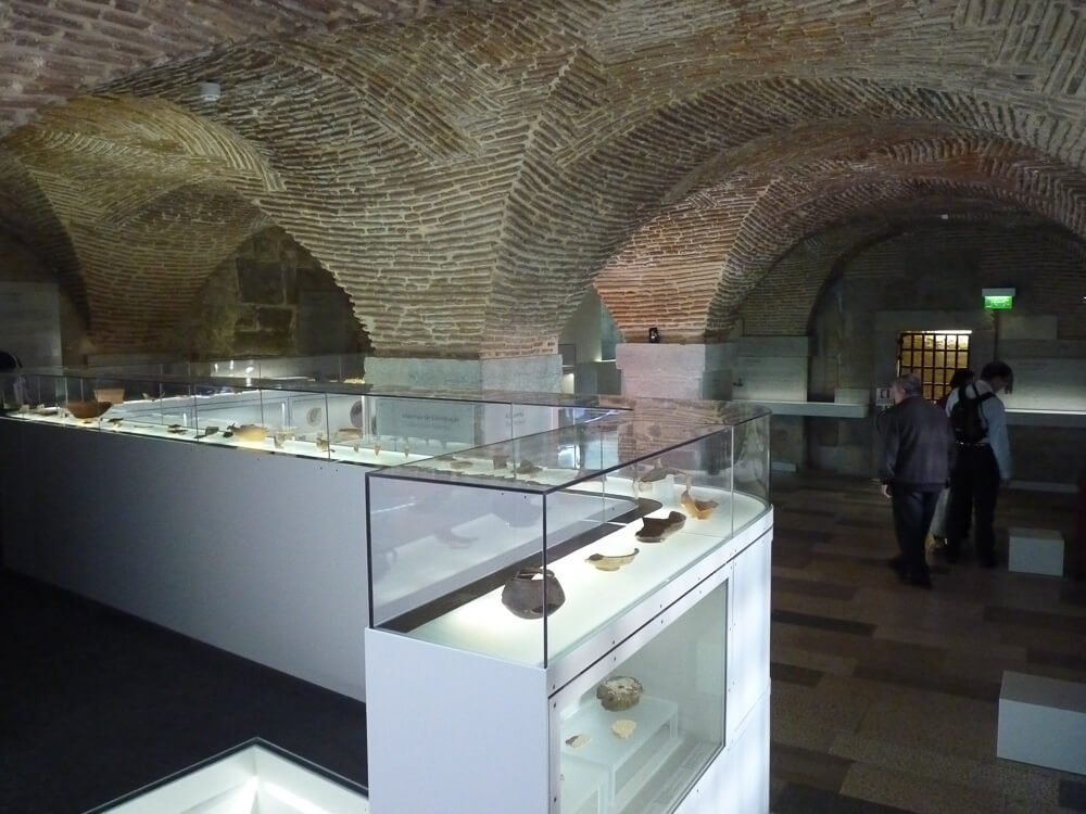 The interior of an exhibition room in a museum with brick arches and display cases showcasing historical artifacts.