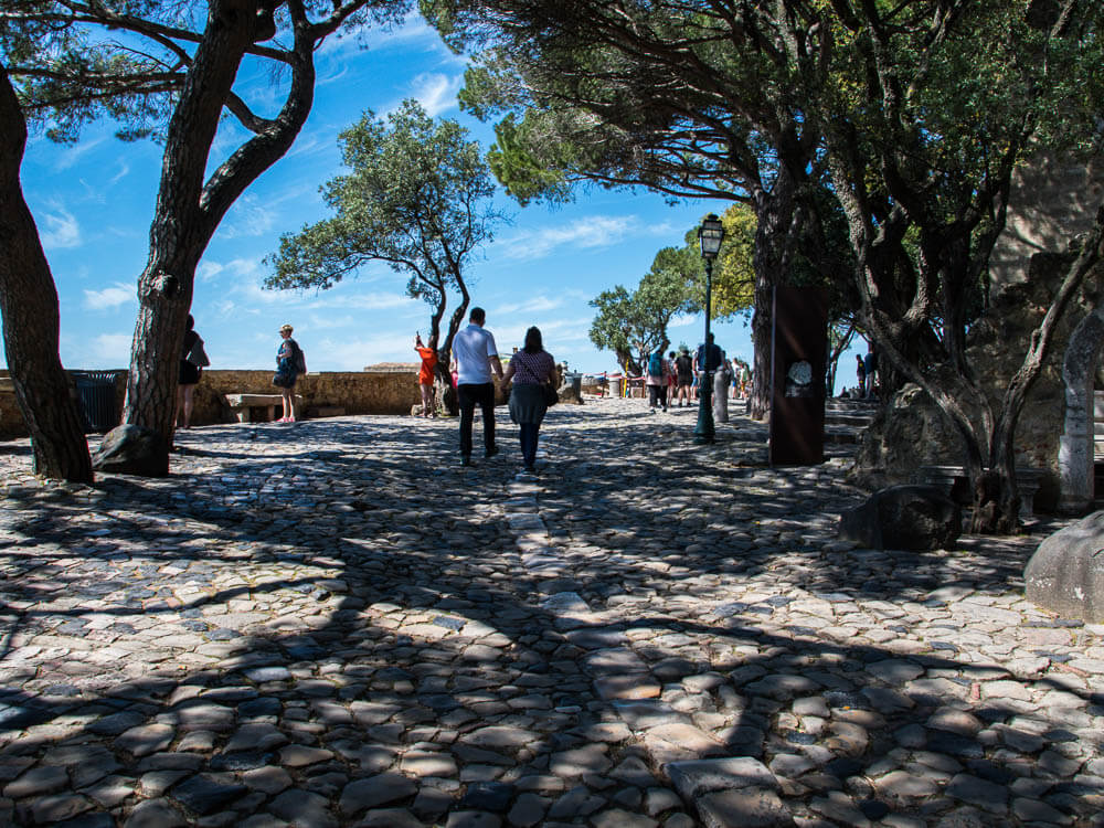 Visitors walk along a cobblestone path shaded by trees within the grounds of Castelo de São Jorge on a sunny day.