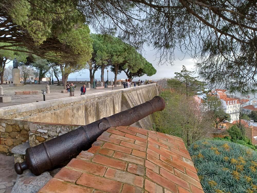 A cannon is mounted on a brick wall overlooking the city and grounds of Castelo de São Jorge, with trees lining the background.