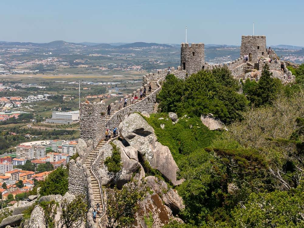 Stone towers and battlements of the Castle of the Moors overlooking a valley with buildings in the distance.