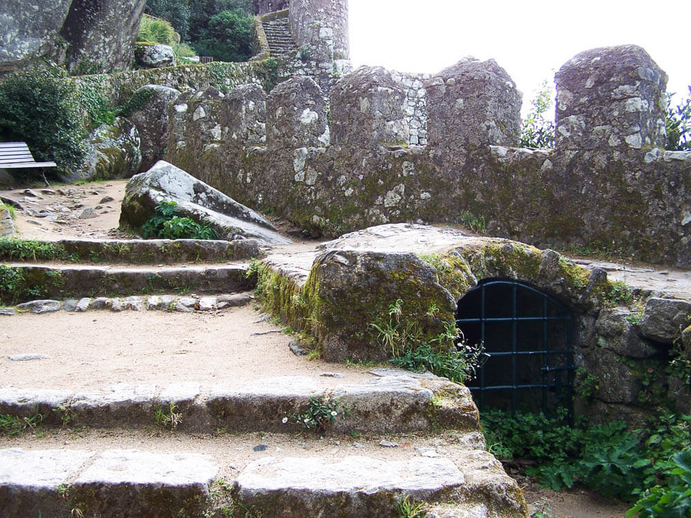 Stone steps leading up to a path along an ancient wall, with a small gated opening on the right.