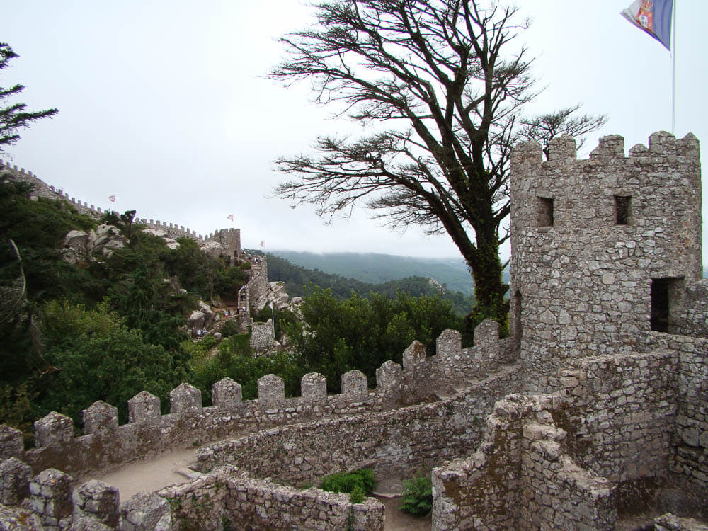 A stone tower and wall with battlements, overlooking a hilly landscape with trees.