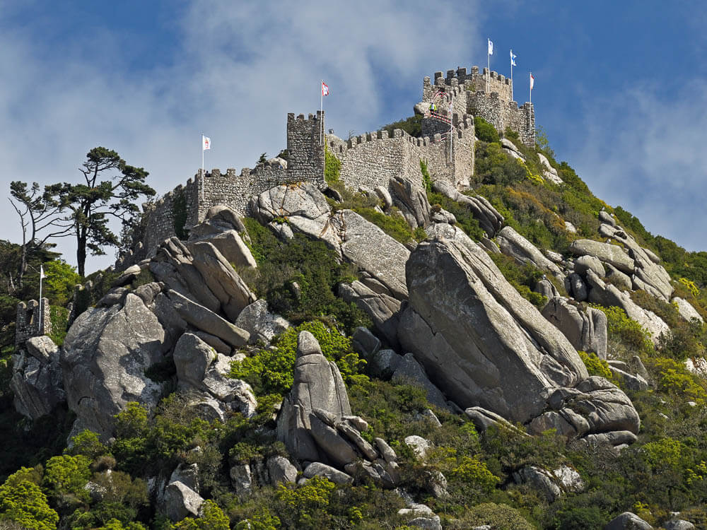 A stone castle perched on a rocky hill, surrounded by greenery and flags flying at the top.