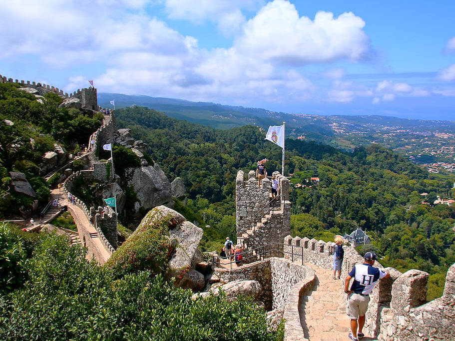 Tourists walking along the stone battlements of the Castle of the Moors, surrounded by lush green forests and expansive views.