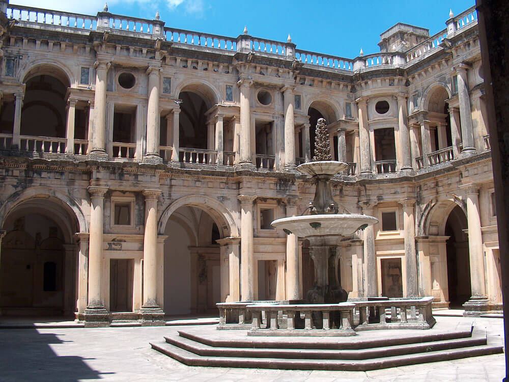 Ornate courtyard of the Convent of Christ in Tomar, featuring a central fountain and a grand, multi-story structure with columns and arches.