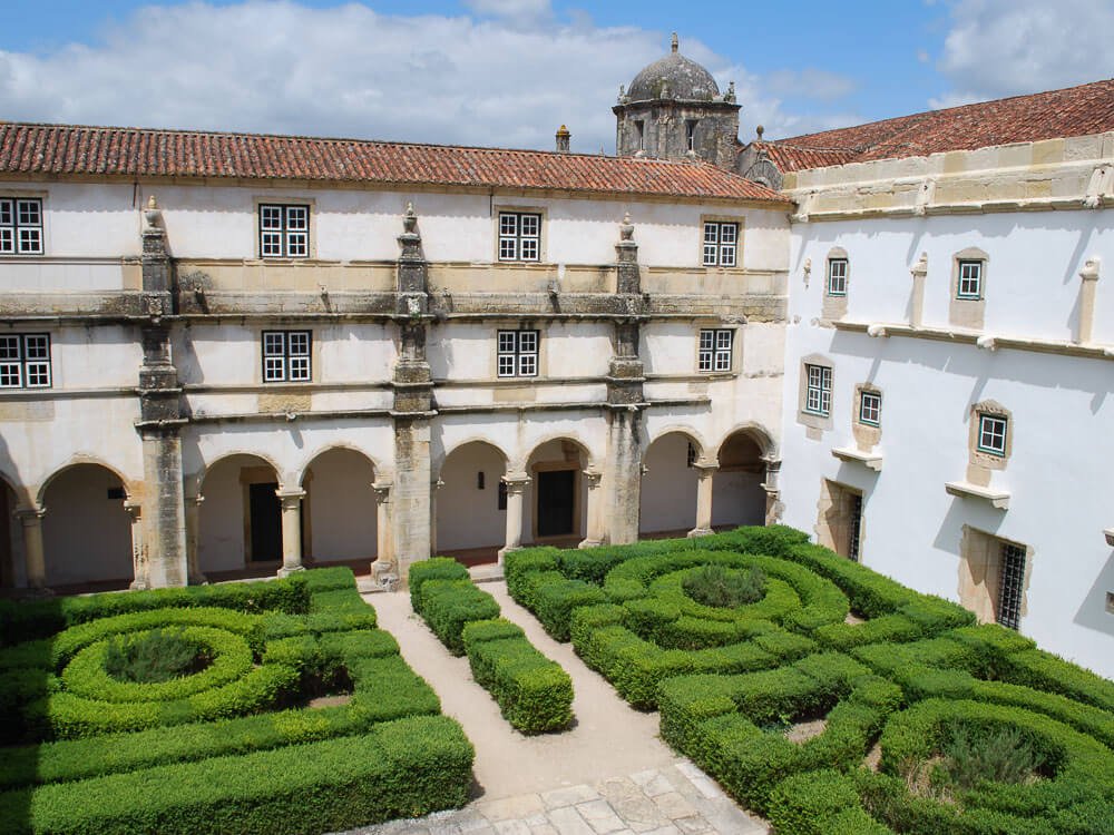 A well-maintained garden with hedges in a cloister, surrounded by white-walled buildings with arched walkways.