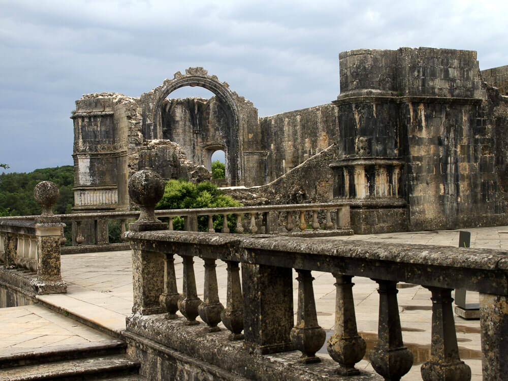 Ruins of an ancient stone structure with a large, weathered arch and a balustraded terrace, set against a cloudy sky.