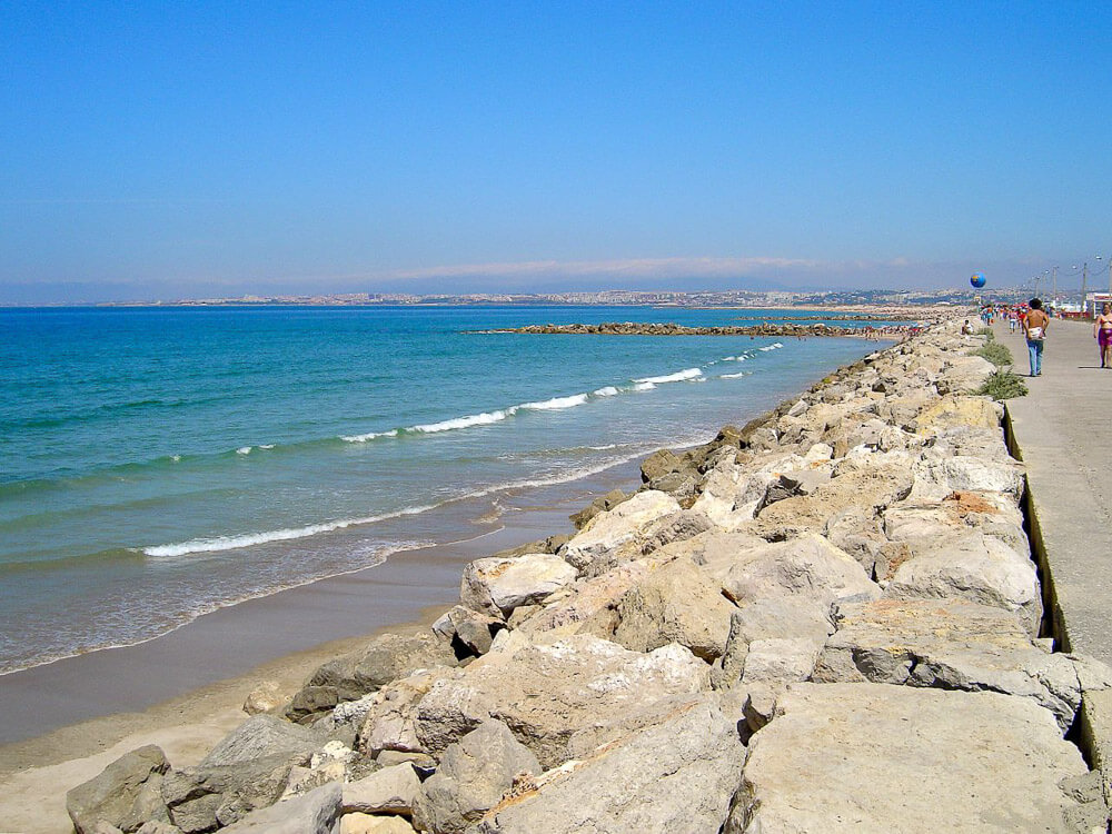 A scenic view of a beach in Costa da Caparica with calm waves, bordered by a rocky breakwater and a promenade.