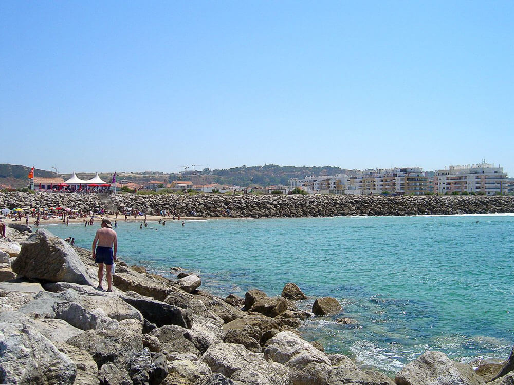 Beachgoers enjoying a sunny day at a beach in Costa da Caparica lined with rocks, with buildings and a beachfront café in the background.