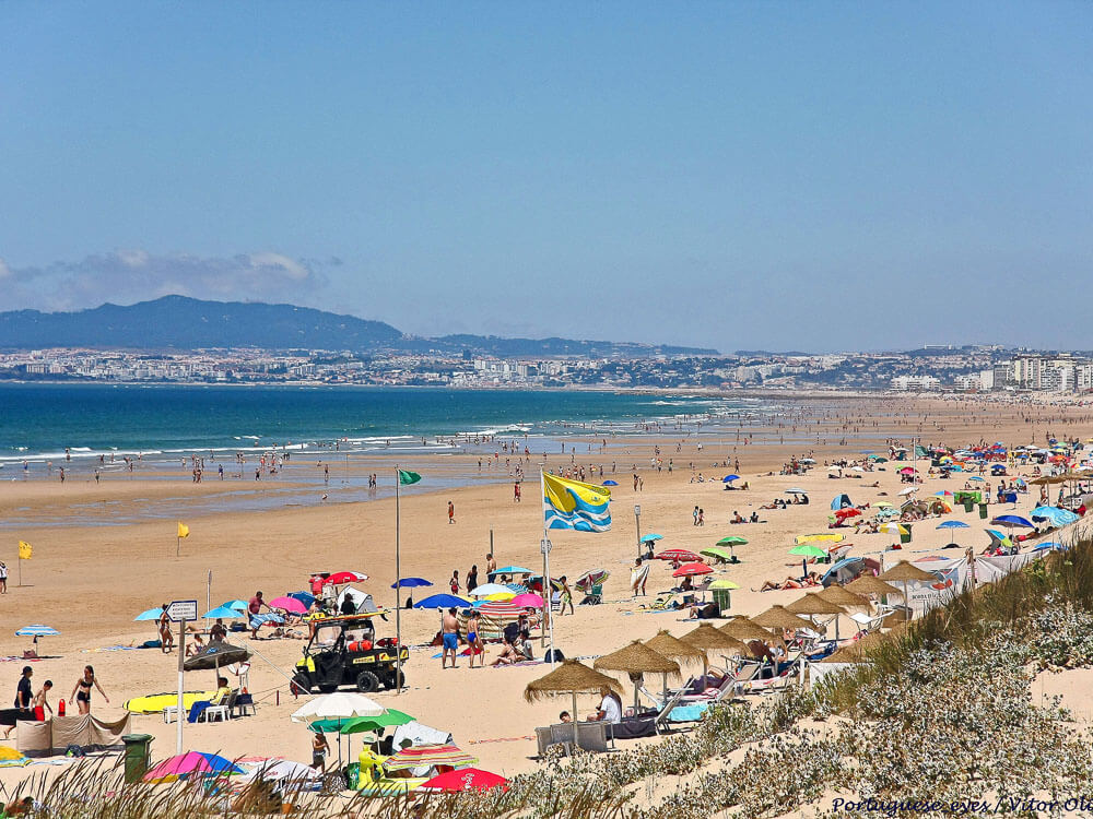 A wide sandy beach crowded with colorful umbrellas and sunbathers, with distant mountains and a city skyline visible in the background.