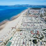 Aerial view of a long beach stretching alongside a town, with rows of beach huts and the ocean extending into the distance.