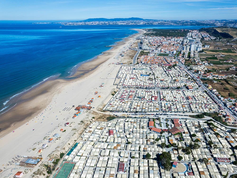 Aerial view of a long beach stretching alongside a town, with rows of beach huts and the ocean extending into the distance.