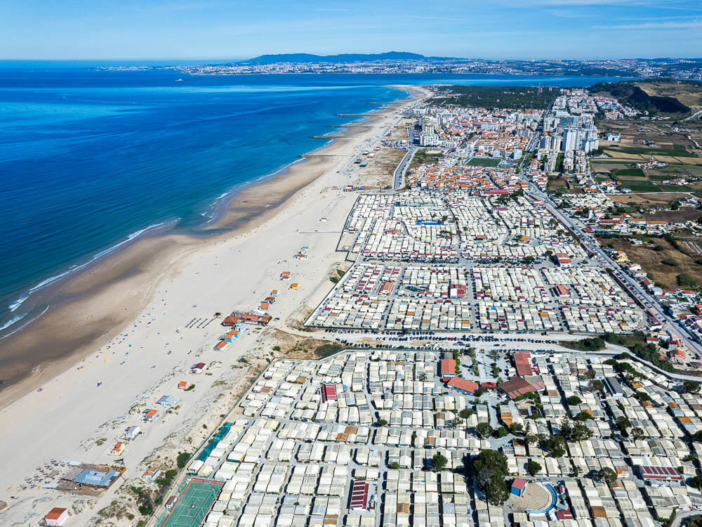 Aerial view of a long beach stretching alongside a town, with rows of beach huts and the ocean extending into the distance.