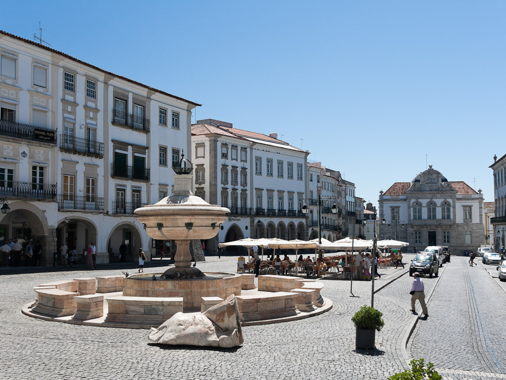 A sunny town square with a large fountain in the center, surrounded by whitewashed buildings with traditional Portuguese architecture.