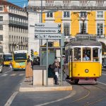 A yellow tram in Lisbon at a city stop, heading towards "Prazeres." People are waiting, with historic buildings and traffic in the background. The signpost points to Rossio, Restauradores, and Santa Apolónia