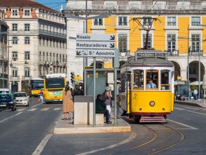 A yellow tram in Lisbon at a city stop, heading towards "Prazeres." People are waiting, with historic buildings and traffic in the background. The signpost points to Rossio, Restauradores, and Santa Apolónia