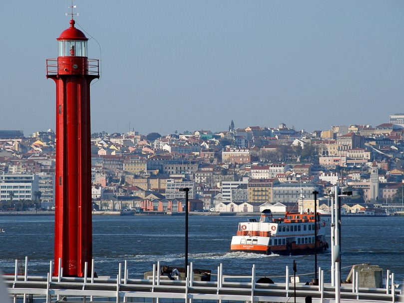 A bright red lighthouse near the water, with a ferry in the background and a cityscape across the river.