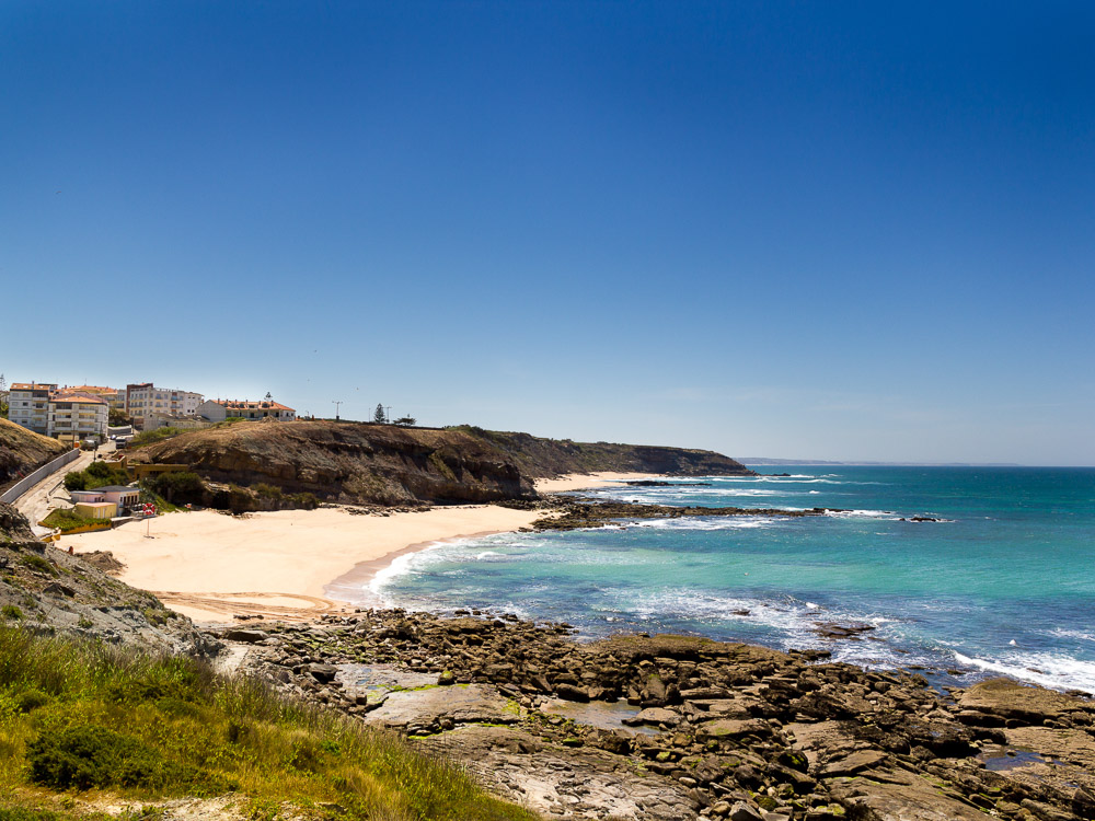 A serene beach with clear blue waters and rocky cliffs, with a small coastal town visible in the background.