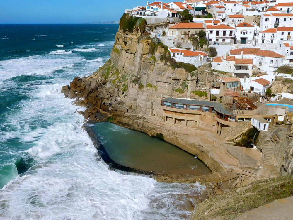 A picturesque coastal village with white houses perched on a cliff, overlooking a natural pool and the rough Atlantic Ocean.