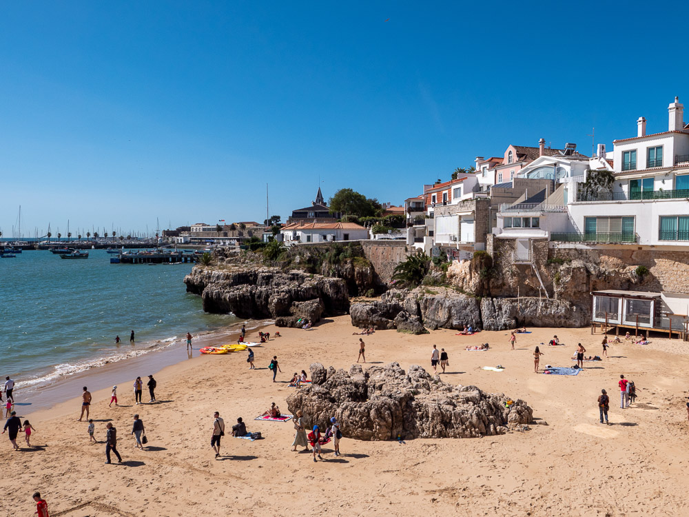 A sandy beach in Cascais with people enjoying the sun, backed by rocky cliffs and seaside buildings.