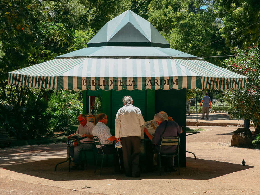 A small green kiosk with a striped canopy and elderly men sitting nearby in Jardim da Estrela.