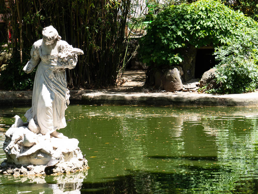 A white marble statue of a woman holding flowers by a pond in Jardim da Estrela park.