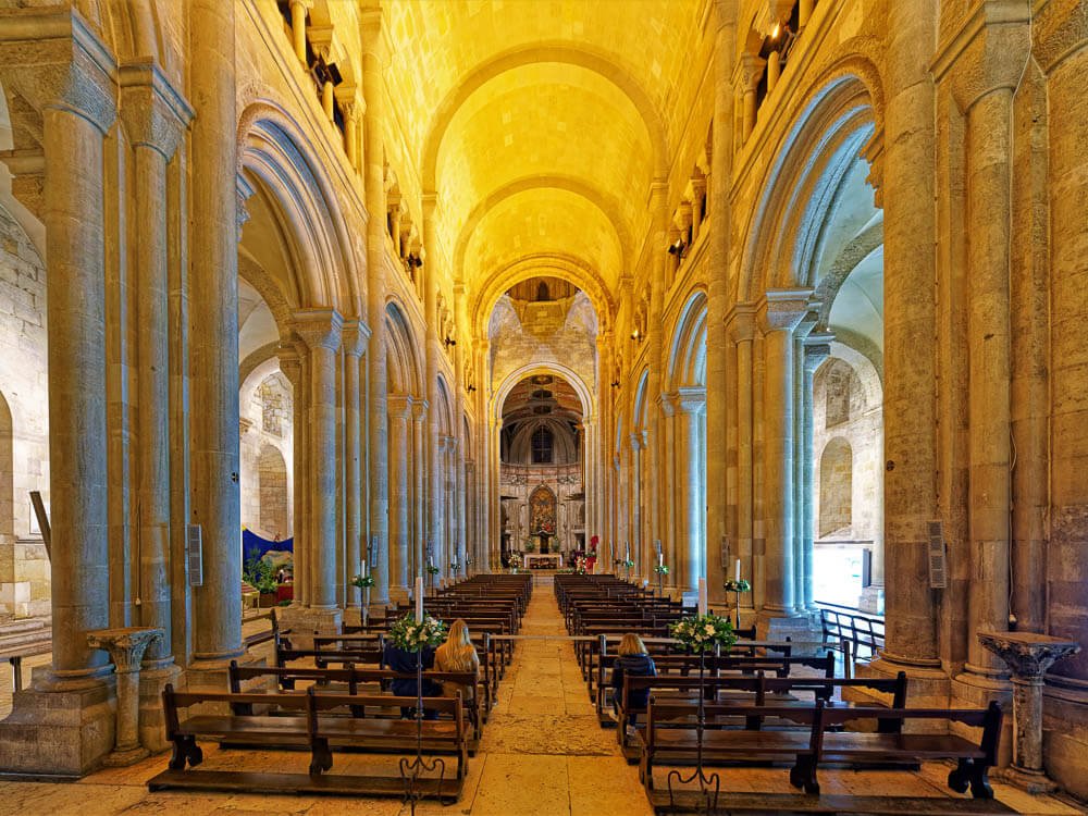 Interior of the Lisbon Cathedral nave with rows of wooden pews and large stone arches.