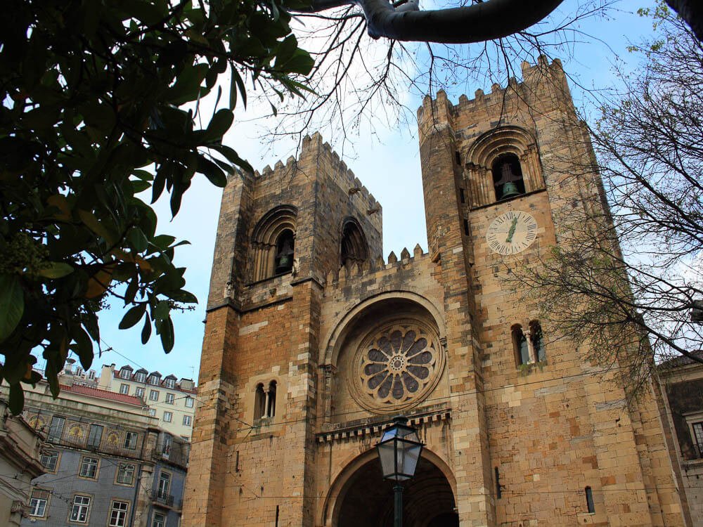 Exterior of the Lisbon Cathedral with two bell towers and a rose window, framed by tree branches.