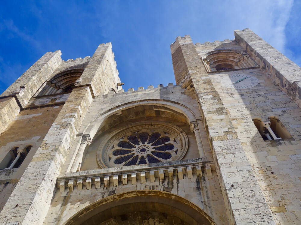 Upward view of the Lisbon Cathedral's façade, showcasing its rose window and twin towers against a blue sky.